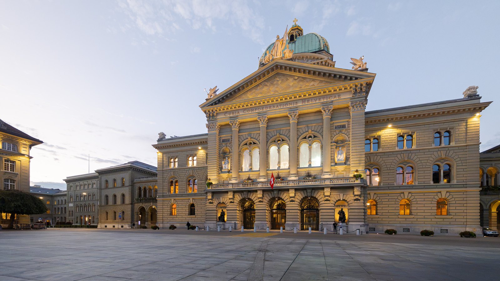 The domed north facade of the Swiss Federal Palace in Bern at dawn, with lit windows and a Swiss flag above the entrance, across an empty stone square