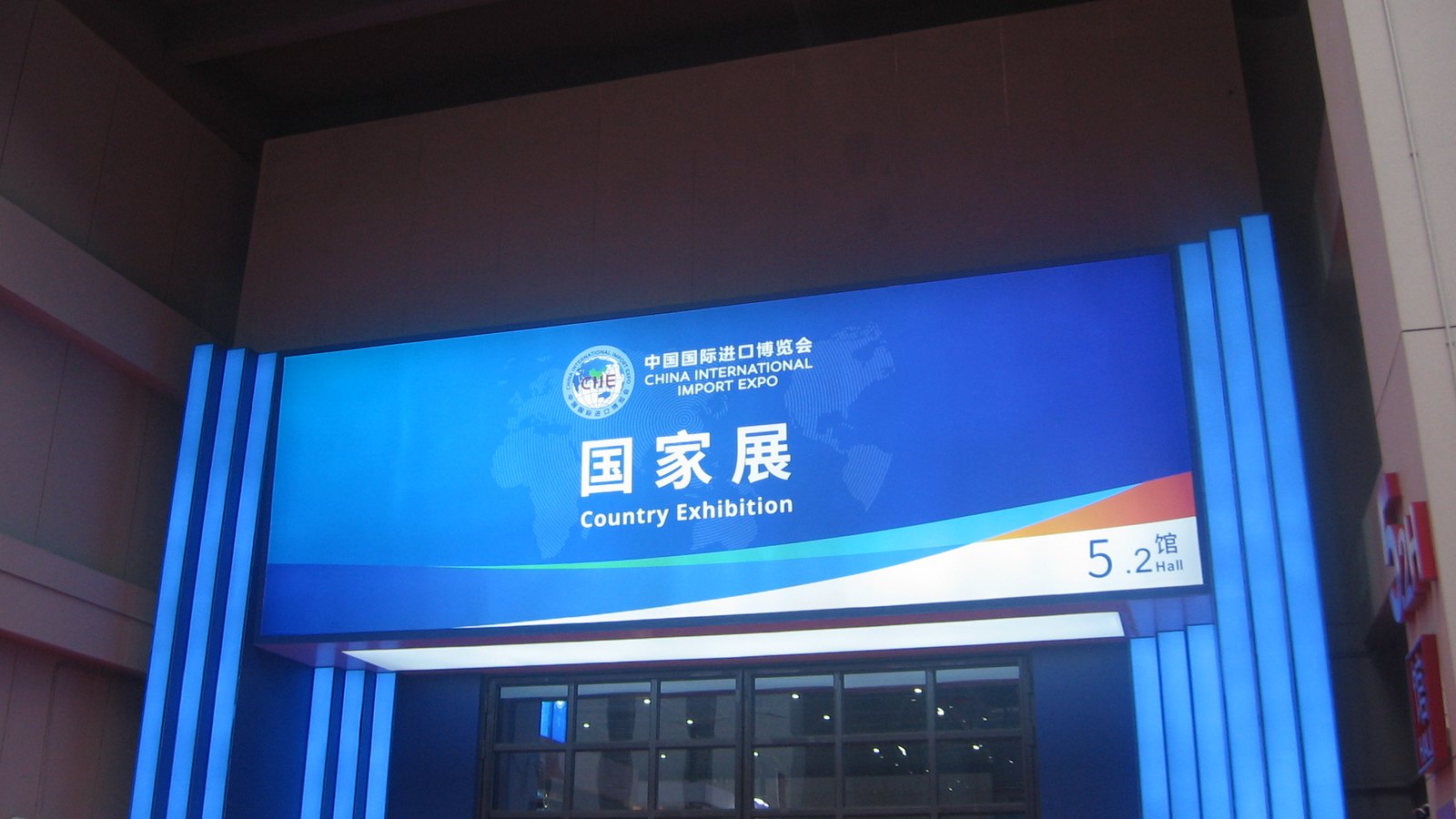 A lit blue sign with the China International Import Expo logo reading Country Exhibition, Hall 5.2, in Chinese and English, above glass entrance doors