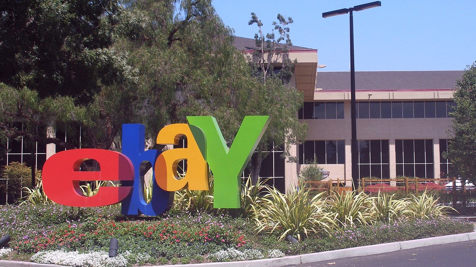 Grandes lettres eBay rouges, bleues, jaunes et vertes dans un massif de fleurs devant un immeuble de bureaux bas