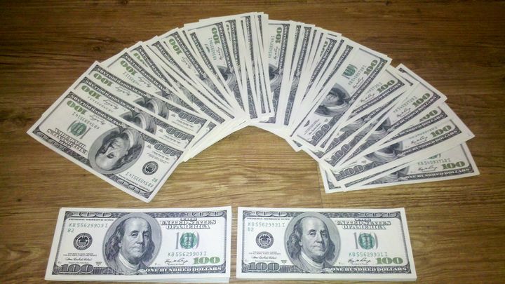 Un éventail de billets américains de cent dollars étalés sur une table en bois, avec deux autres billets posés devant.