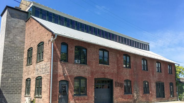 Two-storey red brick building with large windows and a small Etsy sign by the door, under a blue sky.