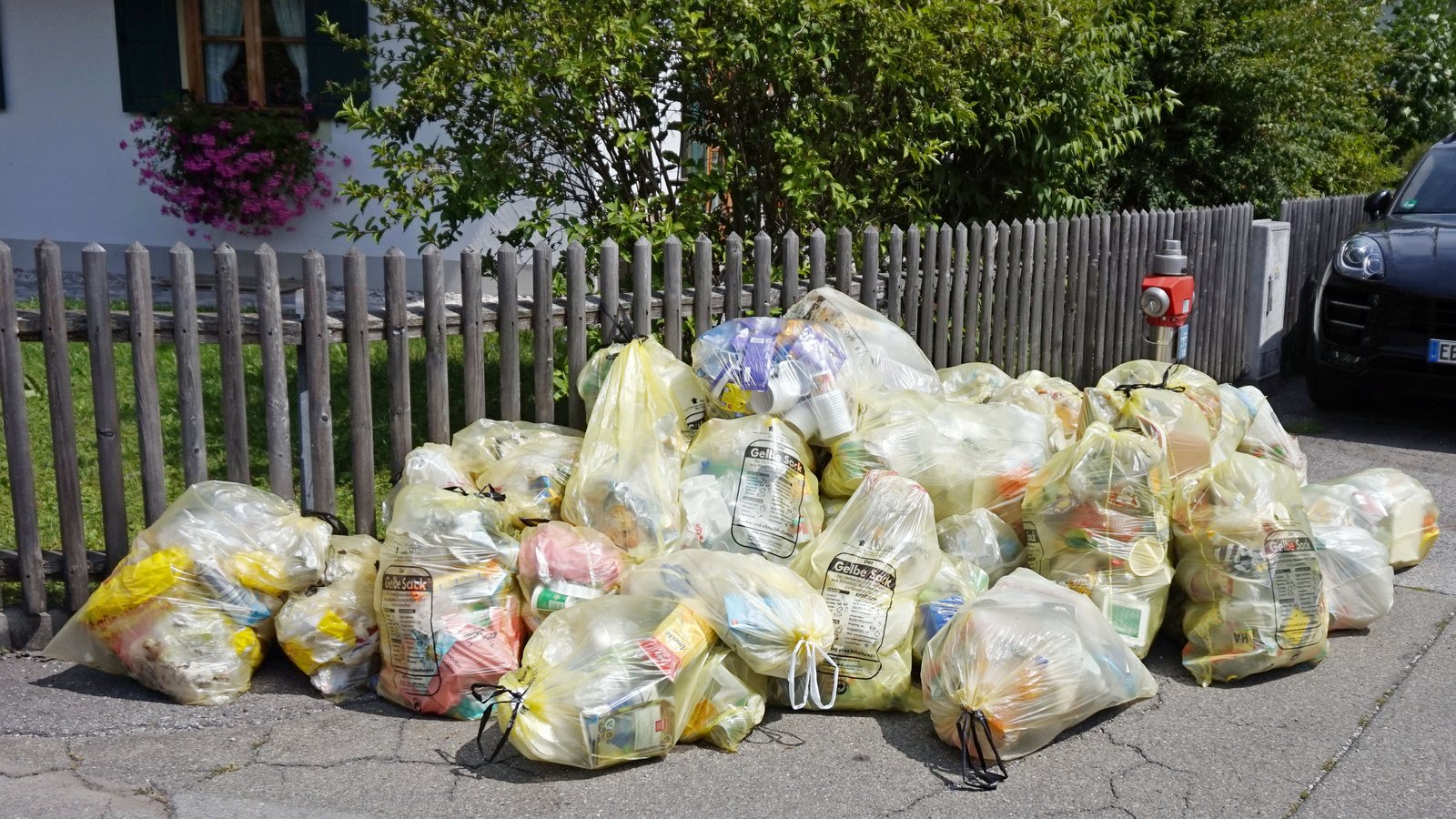 Pile de sacs de recyclage en plastique jaune remplis, posés sur un trottoir devant une clôture en bois et une maison, dans une ville de Bavière