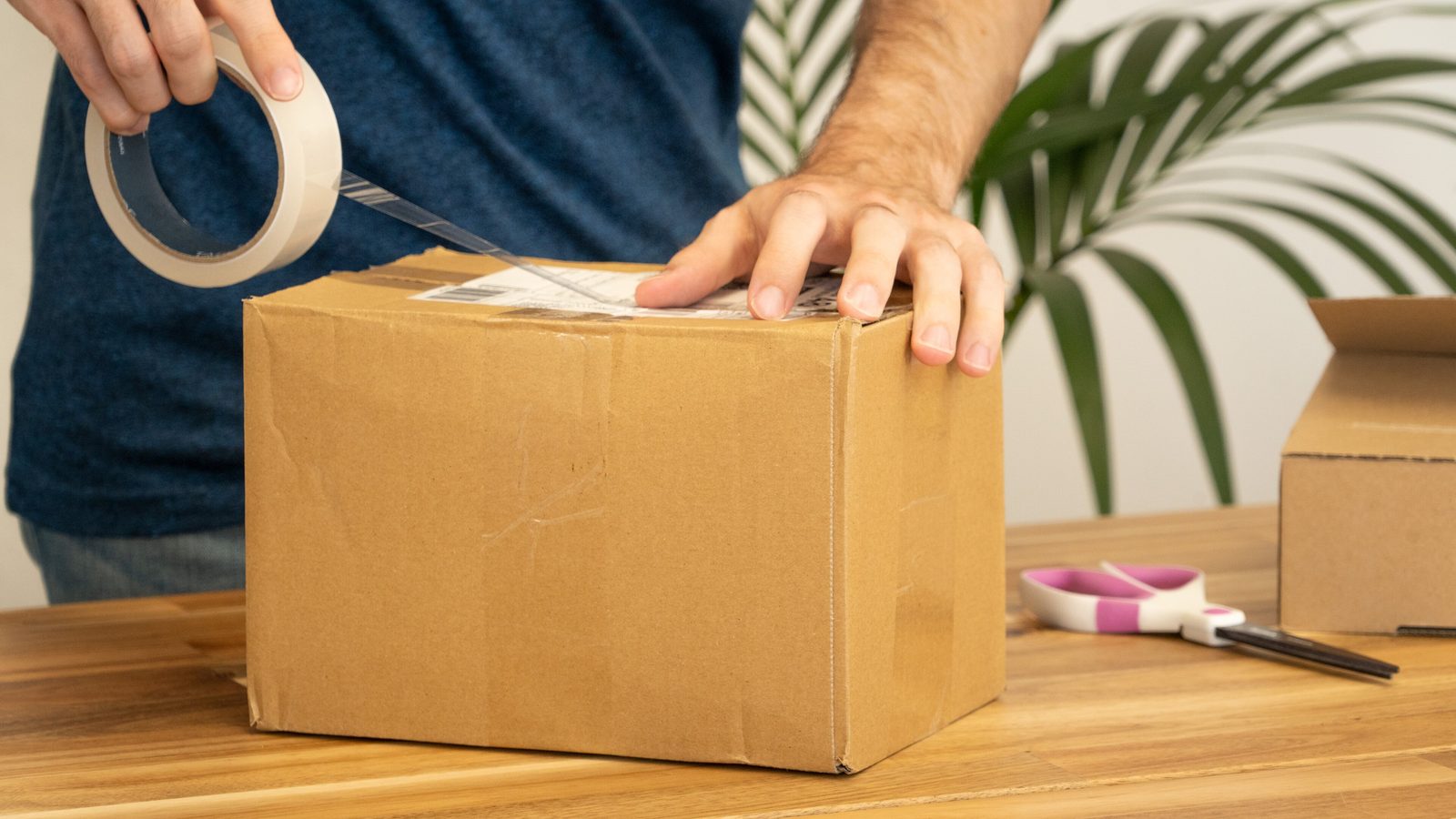 Une personne colle une étiquette d'expédition sur un colis en carton posé sur une table en bois, avec une boîte plus petite et une plante derrière.