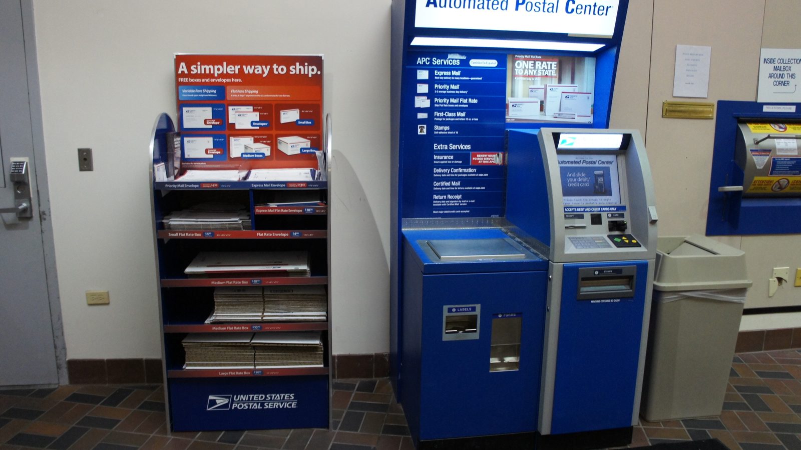 USPS Priority Mail box display next to an Automated Postal Center kiosk in a post office