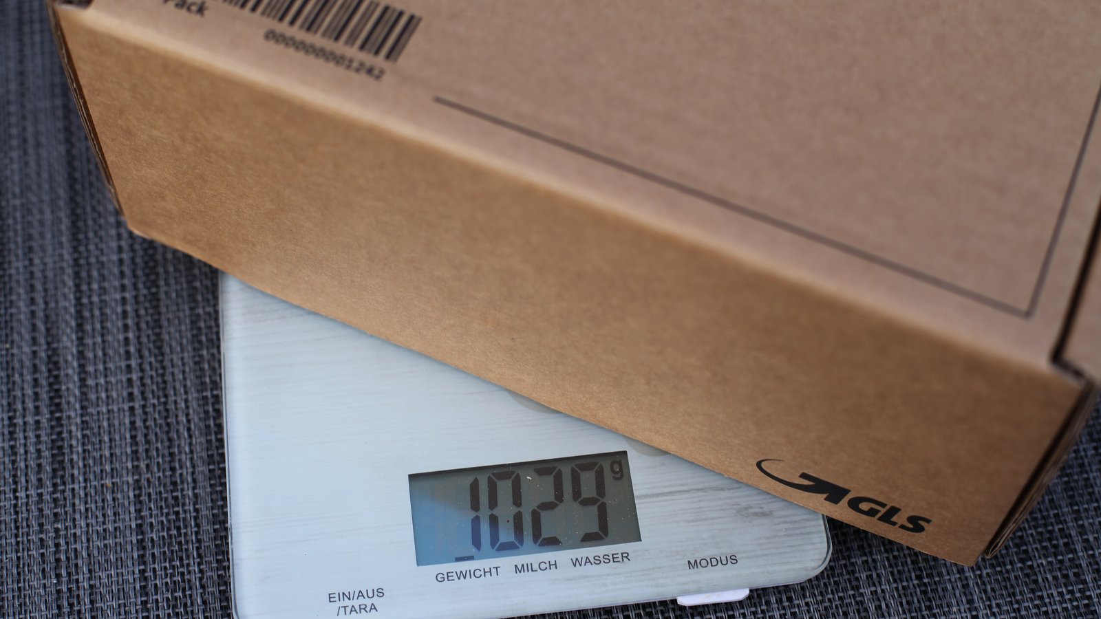 A cardboard shipping box resting on a digital kitchen scale that reads 1029 grams