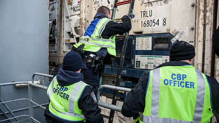 Three CBP officers in yellow vests inspect a cargo container aboard a ship