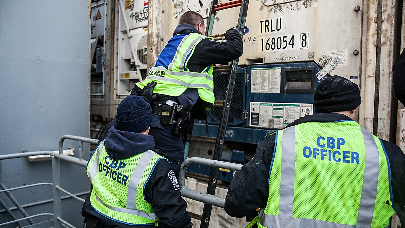 Three CBP officers in yellow vests inspect a cargo container aboard a ship