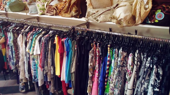 Racks of colorful hanging clothes in a secondhand shop, with handbags lined up on a shelf above.